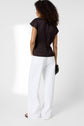 Woman standing with her back to the camera, wearing a top sans manche noir and wide-leg white pants, holding a black clutch in her left hand against a plain light background, accentuated by a subtle bijou doré.