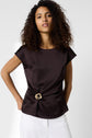 Woman with curly hair wearing a short-sleeved, satin marron blouse featuring a bijou doré decorative buckle at the waist, paired with a white skirt, standing against a plain light background.