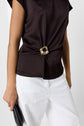 Person wearing a satin marron top sans manche accented with a bijou doré at the waist, paired with white pants and holding a small black handbag.