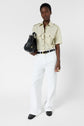 Person wearing a chemise manches courtes beige esprit militaire and wide-leg white pants, holding a black handbag, with a black belt, black shoes, and minimal jewelry, standing against a plain white background.