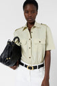 A person wearing a light chemise manches courtes button-up shirt, white pants, and a black belt holds a black handbag. The background is plain and white.