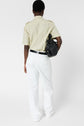 A person stands facing away, wearing a chemise manches courtes beige, white pants, and a black belt, holding a black handbag over their left shoulder, against a plain white background.