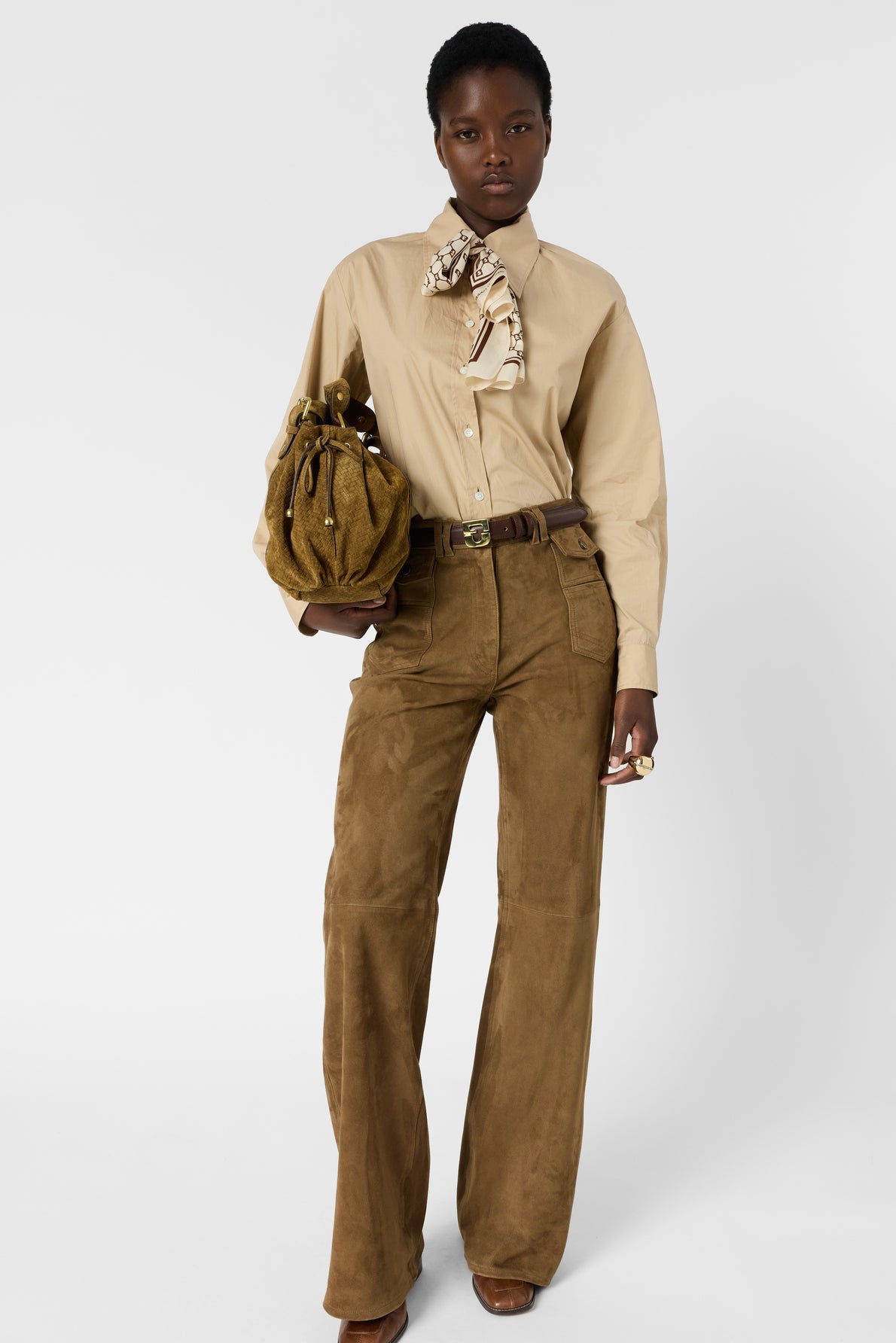 A person stands against a plain background wearing a chemise sable button-up shirt with a scarf, brown wide-leg pants, brown belt, and holding a brown suede handbag. They have short hair and a neutral expression.