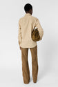 A person stands facing away, wearing a chemise sable and brown suede pants, holding a brown textured drawstring bag under one arm. The background is plain white.
