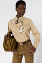 A person wearing a beige chemise popeline with a patterned scarf, brown belted pants, and holding a brown handbag. They have short hair and are posing against a plain light background.