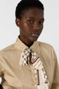A person wearing a beige chemise popeline button-up shirt and a patterned scarf tied at the collar, standing against a plain light background.