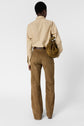 Person standing with their back to the camera, wearing a beige chemise popeline long-sleeve shirt, brown suede pants, and holding a brown suede drawstring bag. The background is plain white.