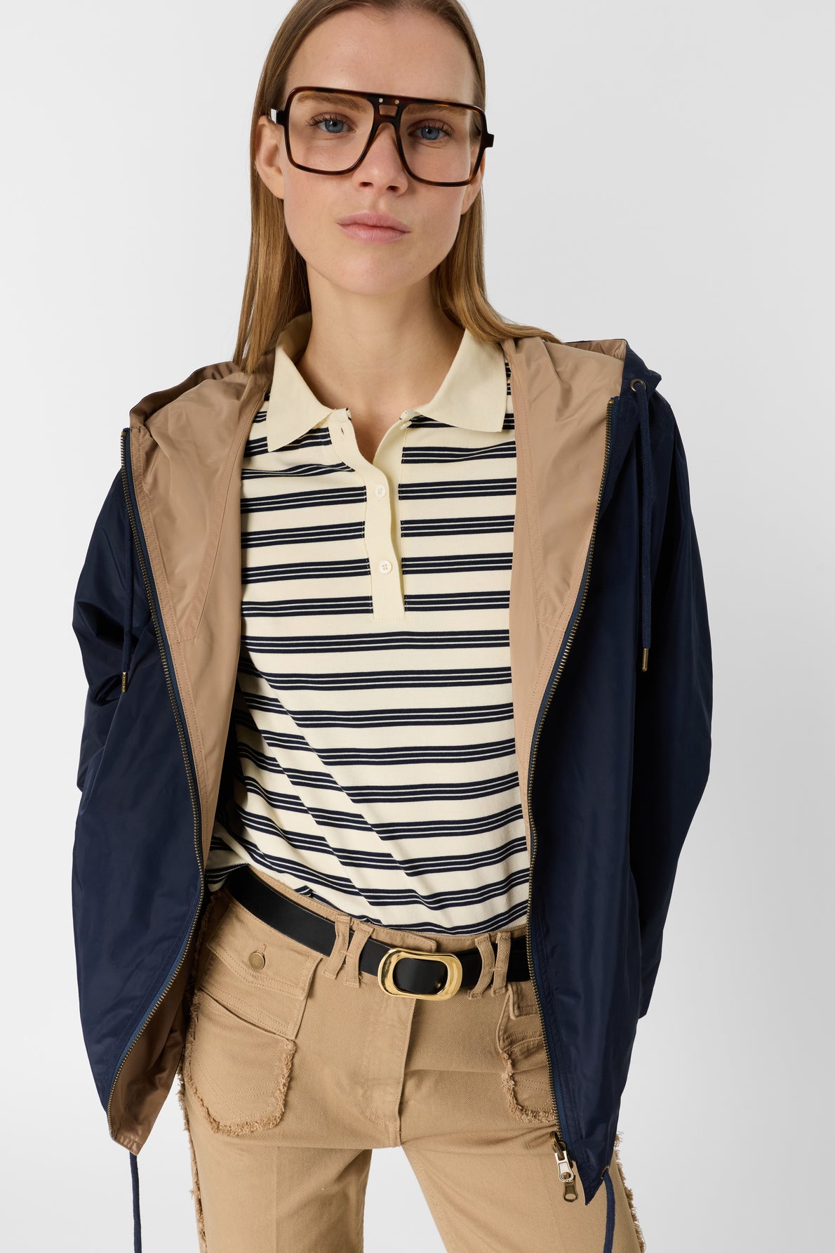 Woman in oversized glasses wearing a beige and black striped polo, tan belted pants, and a navy parka femme with tan lining, standing against a plain white background.