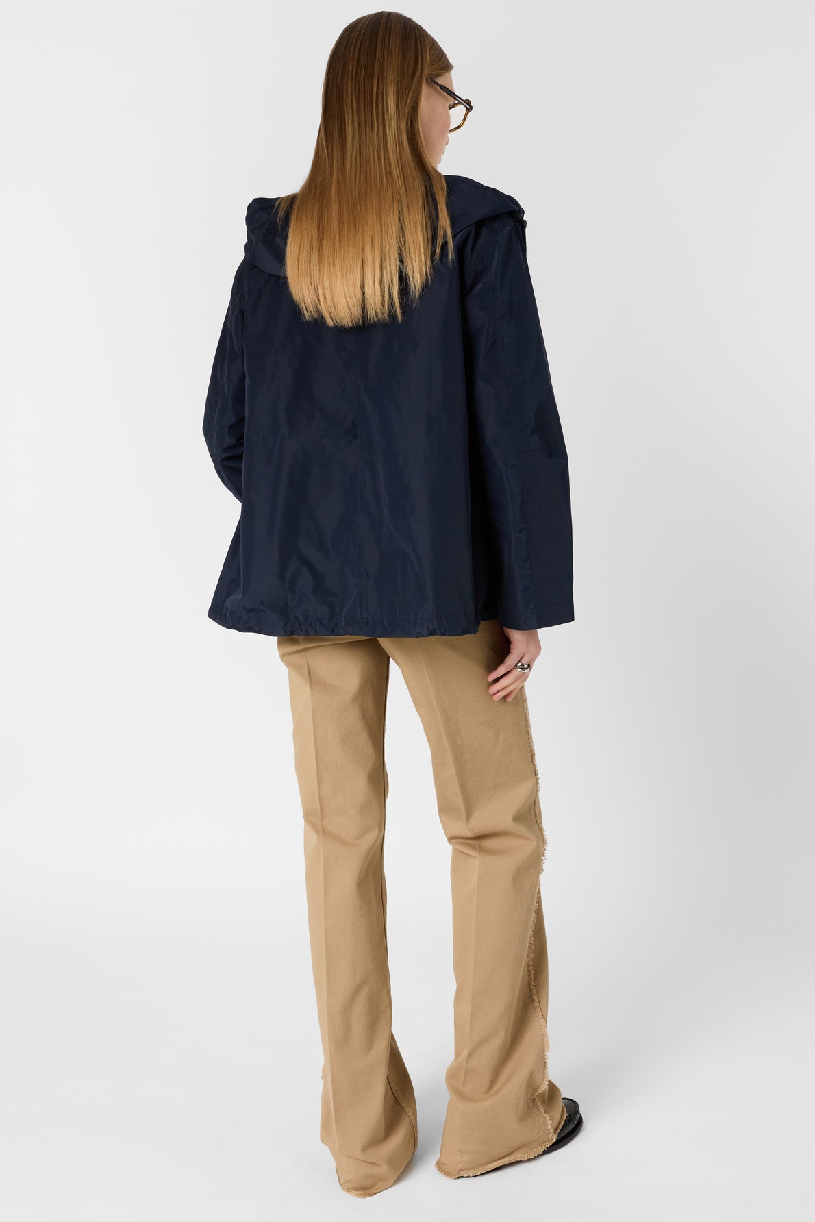 A person with long straight hair wears glasses, a navy parka mi-saison with a hood, and beige pants. They are standing and facing away from the camera on a plain white background.