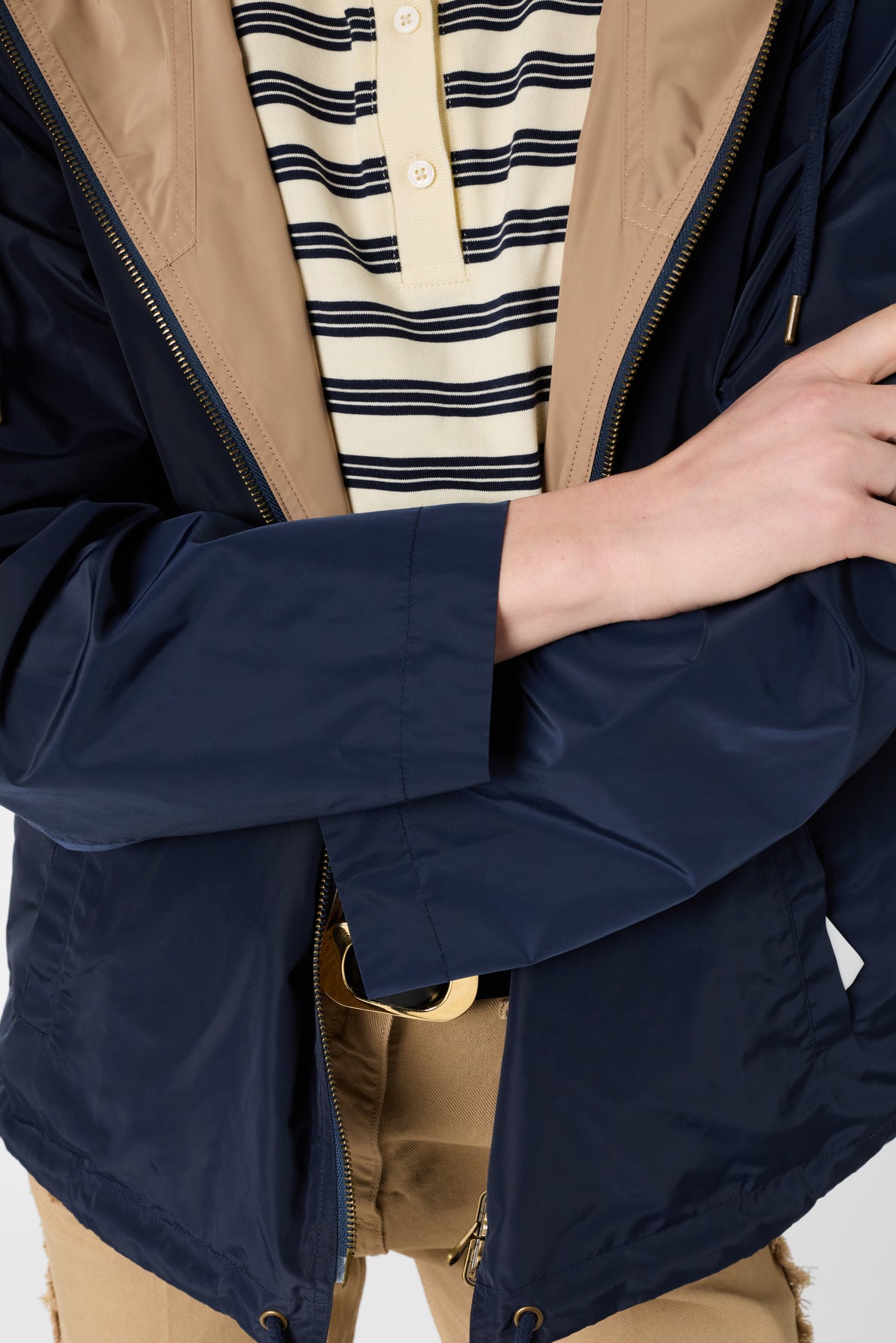 Person wearing a navy parka mi-saison over a beige and navy striped shirt with tan pants and a gold belt buckle. Their right arm is crossed over their body, and their jacket is partially unzipped.