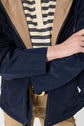 Person wearing a navy parka mi-saison over a beige and navy striped shirt with tan pants and a gold belt buckle. Their right arm is crossed over their body, and their jacket is partially unzipped.