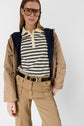 Woman wearing oversized glasses, a beige parka mi-saison with a navy hood, a striped beige and navy polo shirt, and tan pants with a black belt and gold buckle, standing with one hand in her pocket.