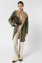 A person wearing a two-tone green and tan parka mi-longue over a tan shirt and cream pants, holding a green handbag, stands against a plain white background.