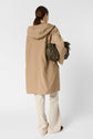 A person with long brown hair, seen from behind, wears a tan veste mi-saison à capuche, cream pants, and holds an olive green purse. The background is plain white.