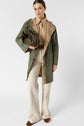A woman wearing a green parka mi-longue hooded coat, beige shirt, cream pants, and brown shoes holds a matching green handbag against a plain white background.