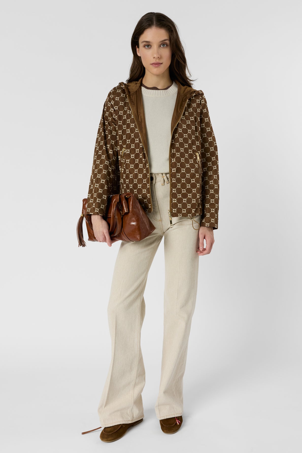 Woman standing against a plain background, wearing a brown patterned veste coupe-vent over a cream sweater, cream pants, and brown shoes, holding a brown leather handbag in her left hand. She has long brown hair and a neutral expression.