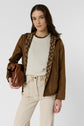 A woman in a brown jacket with a patterned hood, cream sweater, and light beige pants holds a large brown leather purse. She stands against a plain white background, looking at the camera with a neutral expression. Her ensemble is elevated by the addition of a chic parka courte and subtle monogramme écru details that add sophistication to her look.