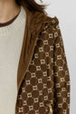 A person wearing a cream sweater with a brown collar and a brown parka courte jacket featuring a beige geometric GG pattern and a gold zipper. Only part of the upper body is visible.