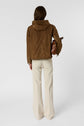A person stands facing away, wearing a brown veste coupe-vent and cream-colored pants. They hold a brown handbag in their left hand against a plain light background.