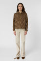 A woman stands facing forward, wearing a brown patterned veste coupe-vent, cream-colored pants, and brown shoes against a plain white background.