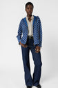 A person stands wearing an édition limitée blue patterned veste coupe-vent, a white patterned blouse, dark flared jeans, a black belt, and black shoes. They have short hair and accessorize with rings and gold necklaces against a plain background.