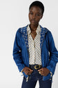 A woman wearing a blue veste coupe-vent with a patterned shirt, dark jeans, and a gold chain necklace stands facing forward with her hands holding the jacket’s zipper.