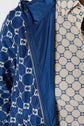 A close-up of a person wearing a blue veste coupe-vent with a white geometric pattern and a beige shirt with a similar blue pattern. Both garments, part of an édition limitée collection, feature repeating chain-like designs.