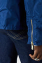 A person wearing a blue veste coupe-vent with a gold zipper and dark blue jeans with white stitching; their hand, adorned with silver rings, rests by their side.