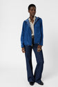 Person wearing a blue veste coupe-vent hooded jacket over a patterned shirt, blue flared jeans, black shoes, and rings, standing against a plain white background.
