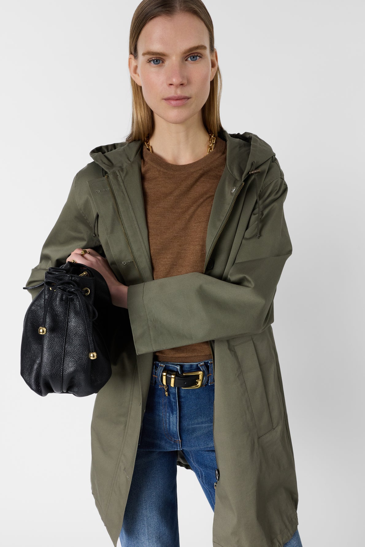A woman wearing a khaki green coat, brown shirt, and blue jeans holds a black leather purse with gold accents, standing against a plain light background.
