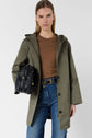 A woman wearing a khaki hooded coat, brown top, and blue jeans holds a black handbag. She stands against a plain light background, gazing confidently at the camera.