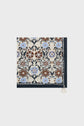 A carré en soie featuring an intricate floral pattern in blue, brown, and cream tones, this accessory bohème is both elegant and versatile. The square scarf boasts a symmetrical imprimé de rosaces design with a dark border and a white tassel attached to one corner, creating a decorative flourish that completes any look.