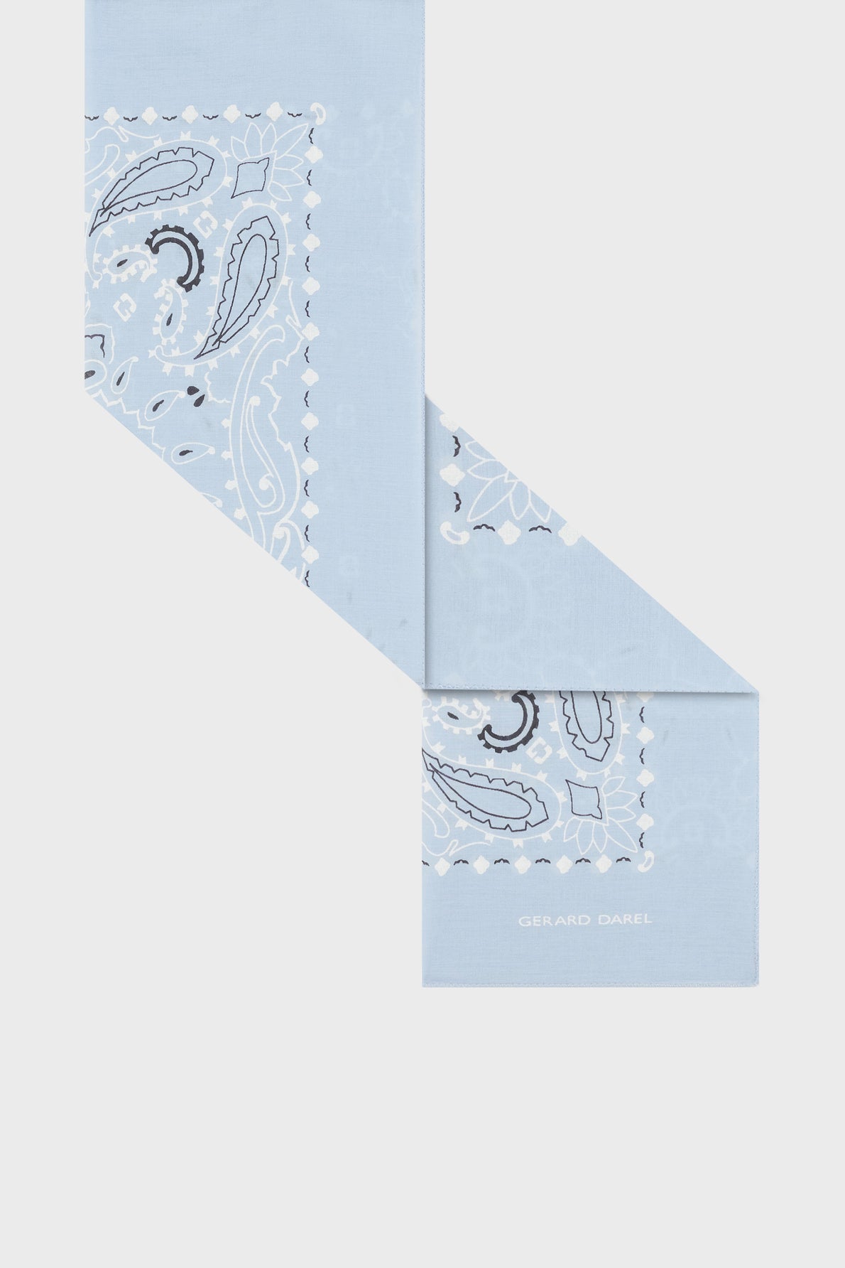 A light blue foulard coton with a white and black paisley imprimé bandana pattern, folded in a zigzag shape. The corner displays the text GERARD DAREL in white letters, reminiscent of the iconic carré Dayana style.