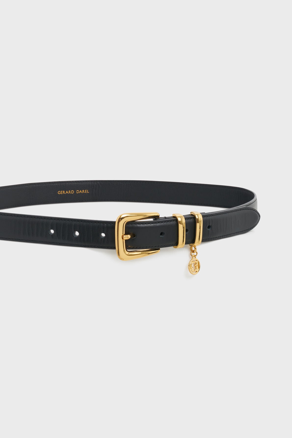 A black leather belt with a gold buckle and a small gold charm attached, displayed on a plain white background. The brand name GERARD DAREL is printed in gold inside the belt, highlighting this ceinture élégante designed for the modern woman.