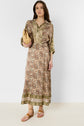 Woman wearing a long patterned dress with green and brown motif floral designs, holding a woven clutch, and wearing tan sandals, standing against a plain white background.