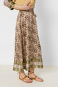 A person wearing a jupe longue with intricate paisley designs and a green floral border, crafted from soft soie, paired with brown strappy sandals. Only the lower half of the body is visible.