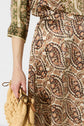 A person wearing a patterned, paisley-print outfit with subtle motif floral details holds a woven, beige clutch bag. Only the lower torso and one arm are visible against a plain background.