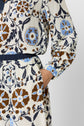 A person in a cream dress with brown and blue floral patterns stands with one hand in their pocket. The focus is on the dress’s vibrant motif végétal and the details of its forme évasée at the waist and sleeve.