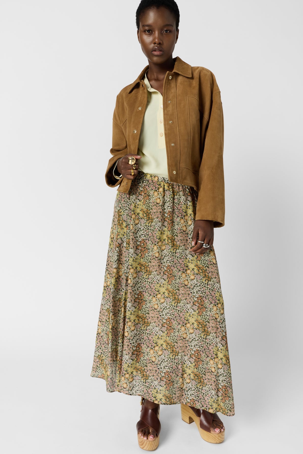 A woman stands wearing a tan suede jacket, a pale yellow top, and a jupe longue à motif floral that embodies un esprit bohème. She completes the look with platform sandals. Her short hair frames her neutral expression as she rests one hand casually in her skirt pocket.