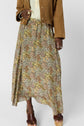 A person wearing a tan suede jacket, cream blouse, and a jupe longue à motif floral in brown, yellow, and green tones embodies un esprit bohème. They are also wearing platform sandals and several rings.