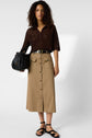 A person stands wearing a brown knit top, beige button-front midi skirt with pockets, black belt, black sandals, and holds a black textured bag. The background is plain white.
