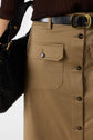 Close-up of a person wearing a brown textured top, a tan button-front skirt with flap pockets, a black belt with a gold buckle, and carrying a black woven handbag.