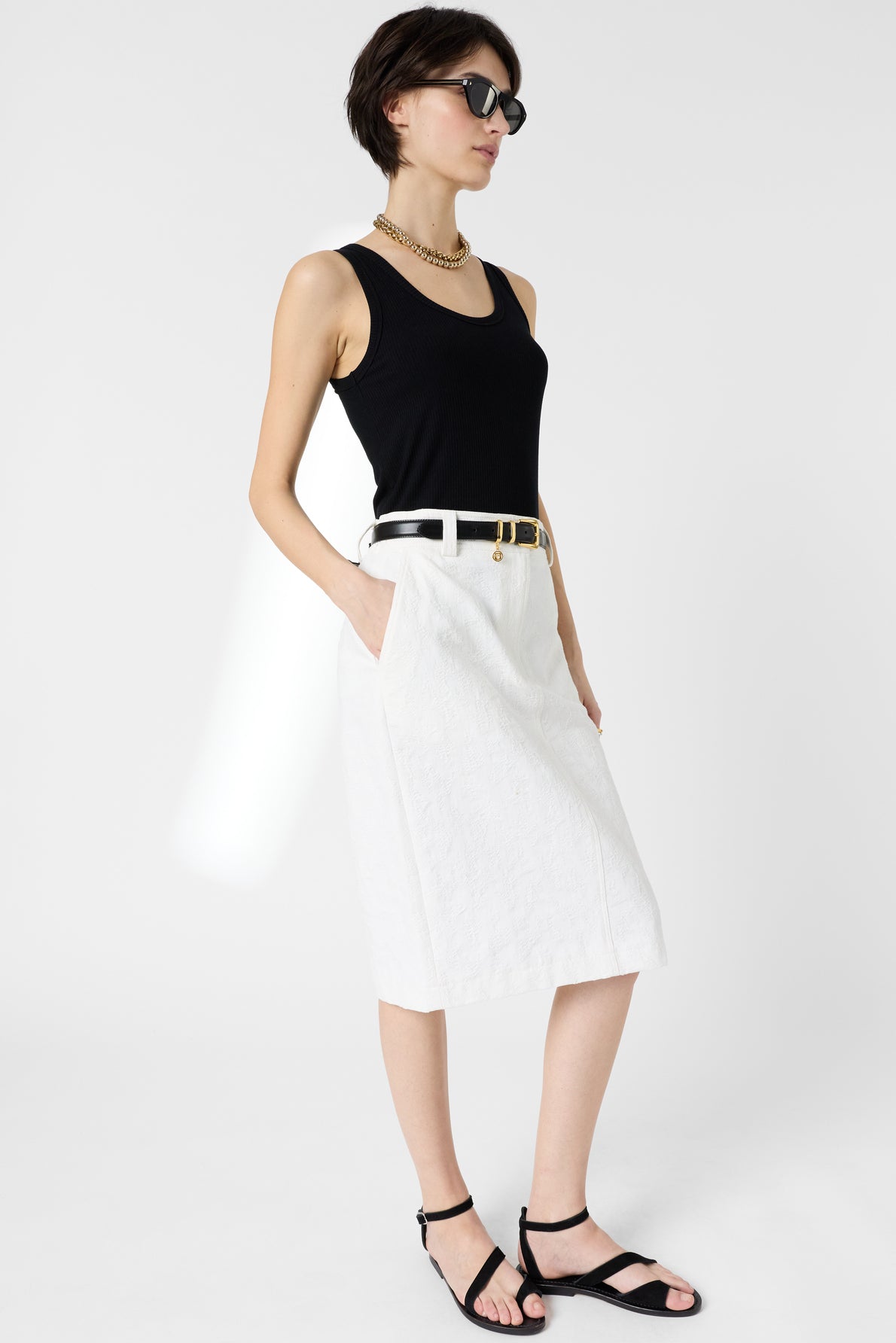 Woman in sunglasses, a black sleeveless top, white knee-length skirt with pockets, black belt, black sandals, and gold necklace stands against a plain white background.