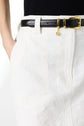 Close-up of a person wearing a white textured skirt, black belt with a gold buckle, and a dangling gold charm. The persons hand is in their pocket and they are wearing a black top.