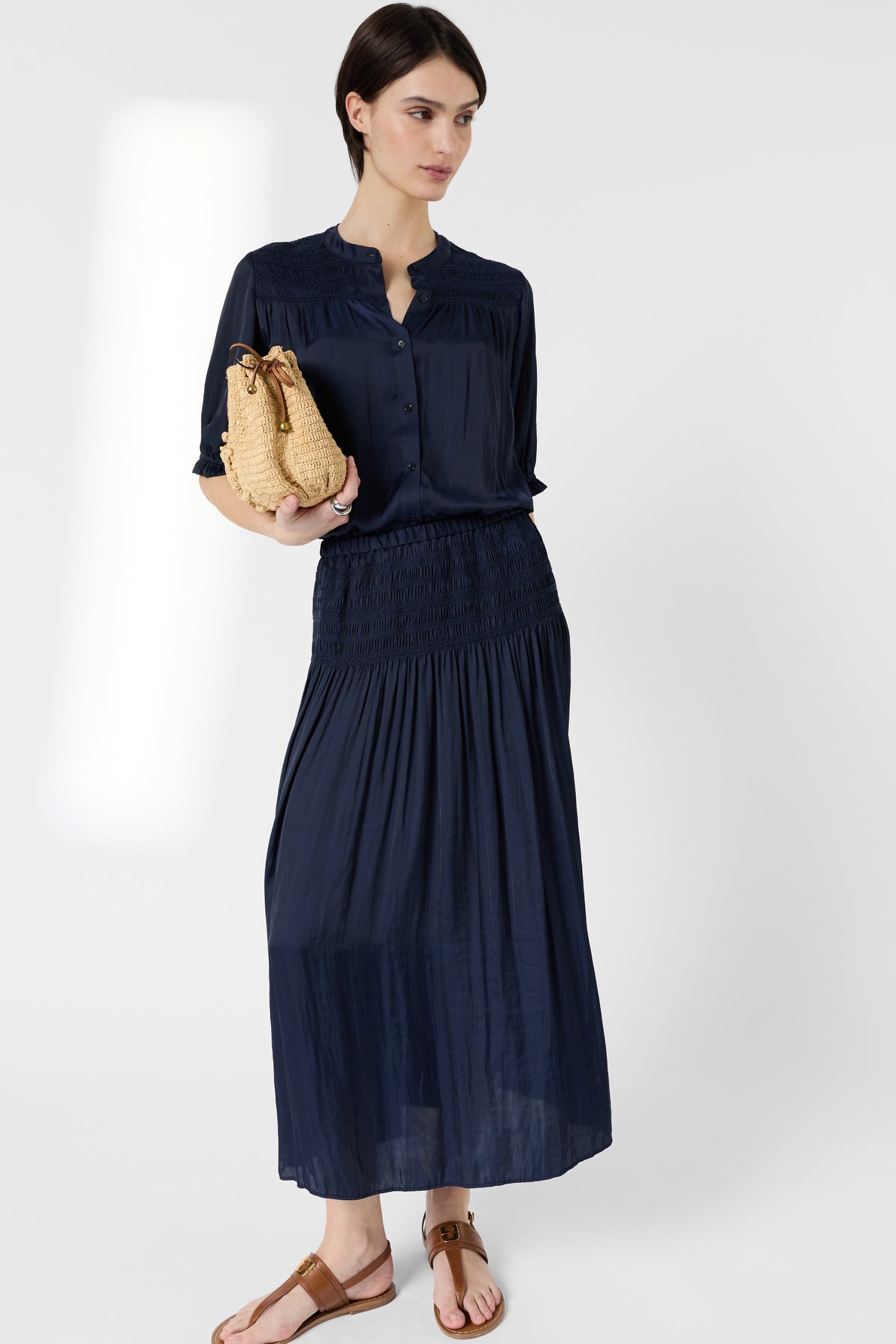 A woman wearing a navy blue button-down dress and brown sandals holds a woven straw handbag, standing against a plain white background—her look effortlessly complemented by the classic charm of a jupe midi style.