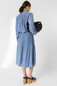 A woman with brown hair in a ponytail wears a light blue, long-sleeve, knee-length dress, black sandals, and holds a black bag, standing against a plain white background, facing away.