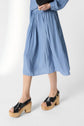 A person wearing a light blue, pleated midi skirt and black leather platform sandals with wooden heels, standing against a plain white background.