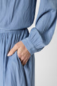A person wearing a light blue, long-sleeve dress with their right hand tucked into the pocket. Only the midsection, arm, and part of the hand are visible against a plain background.