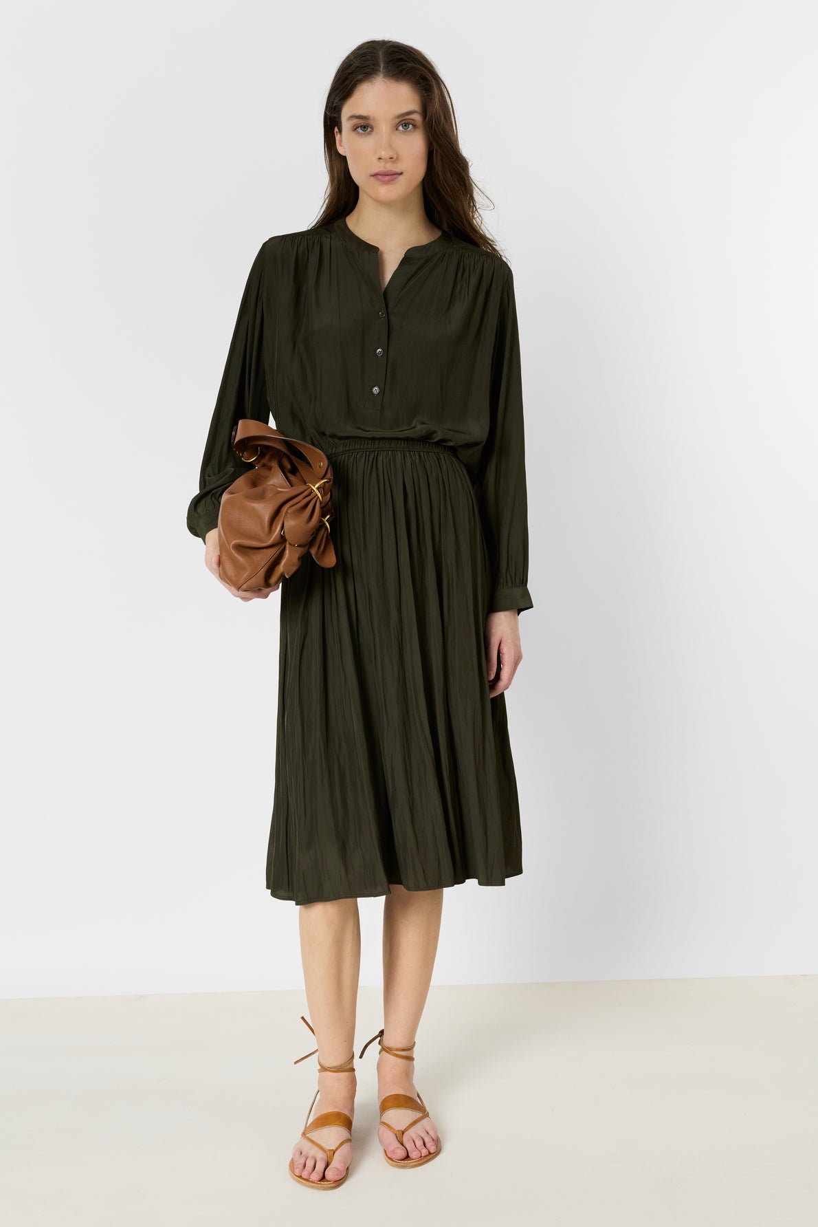 A woman stands against a white background wearing a dark green long-sleeve, knee-length dress, tan sandals, and holding a brown clutch bag.