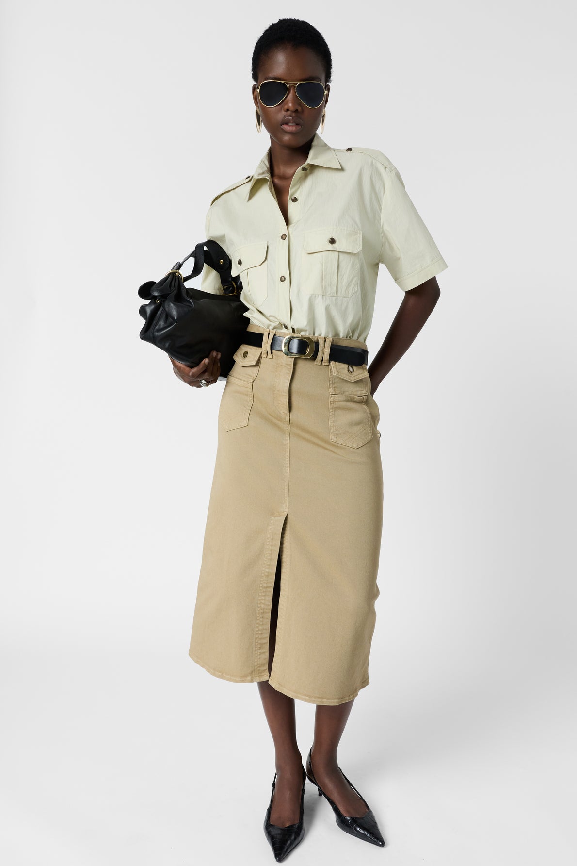 A person wearing sunglasses, a short-sleeve light shirt, a belted tan midi skirt with a front slit, black pointed shoes, and holding a black bag stands against a plain white background.